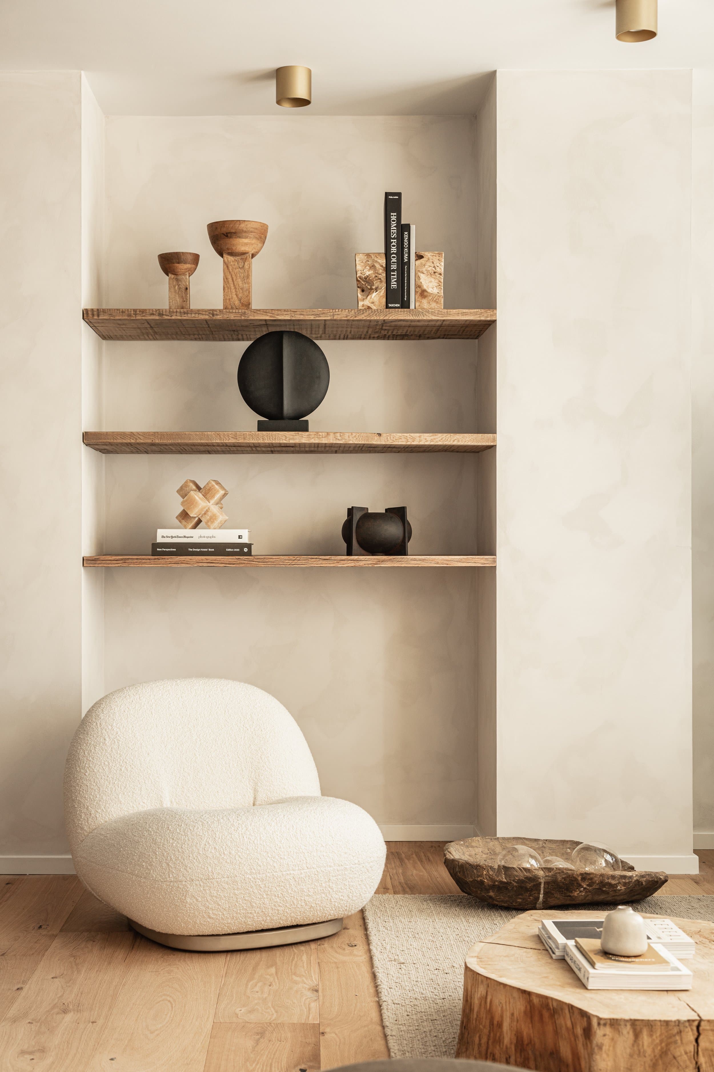 Minimalist living room with wooden shelves displaying decor, a white plush chair, and a wooden coffee table on a light wood floor.